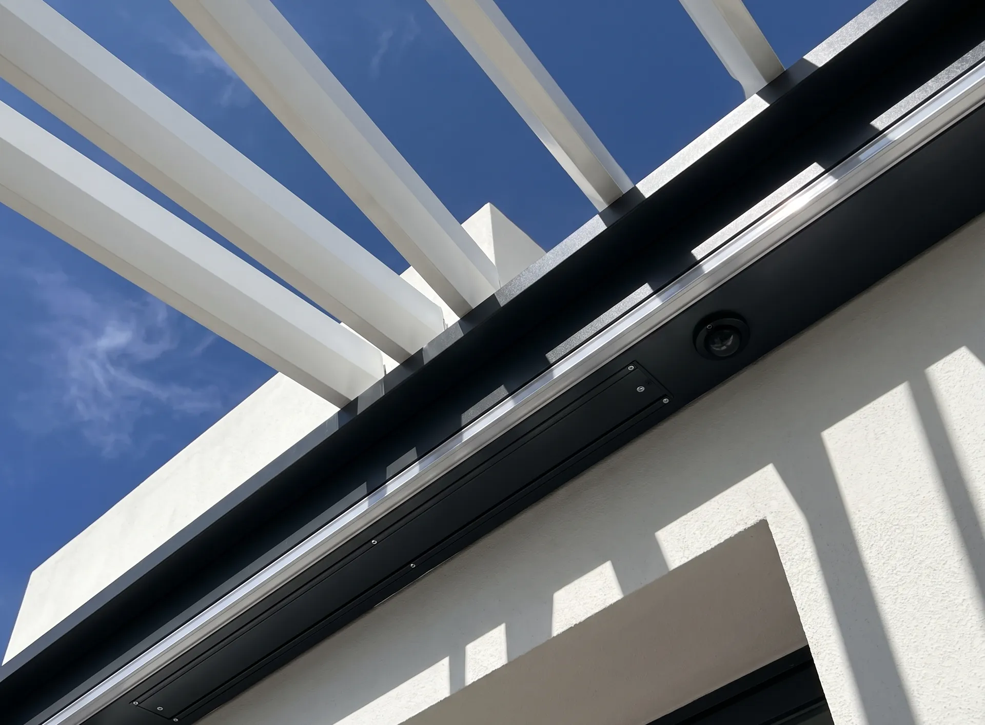 Motorised louvre roof of an aluminium pergola caught mid-rotation, slim drive housing and rain sensor visible on the frame edge