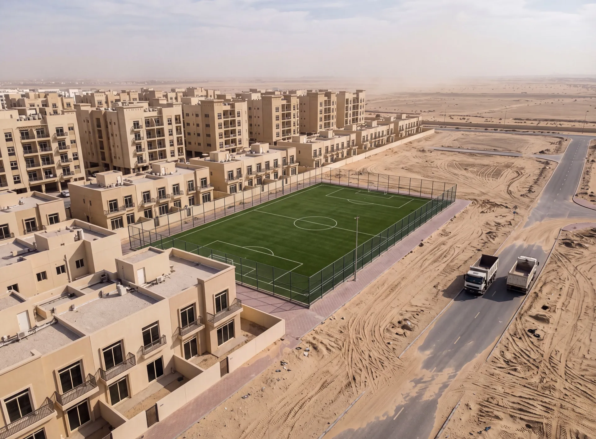 Beige mid-rise apartment blocks and townhouse rows beside a fenced sports pitch and open desert sand in Dubai Sports City